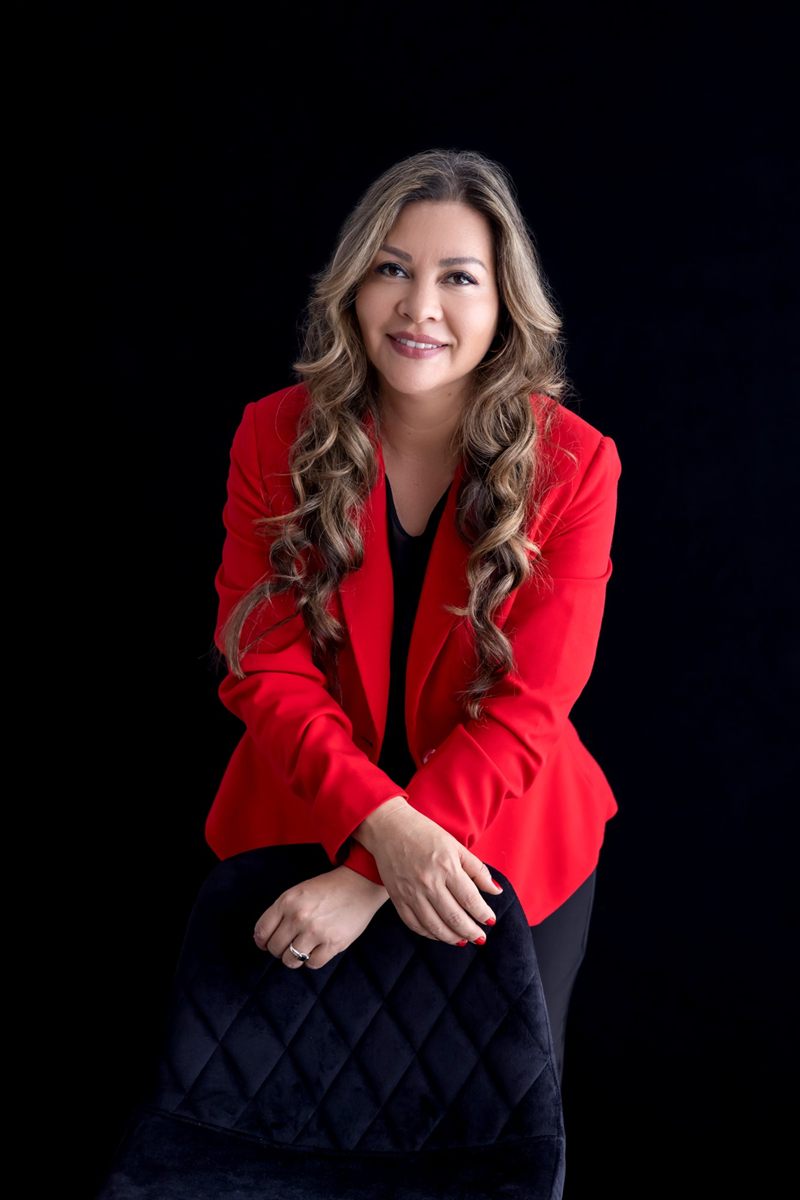Lina Hernández sonríe sobre fondo negro, con blazer rojo, apoyada en una silla negra capitoneada